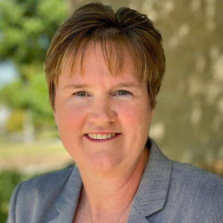 Woman with short hair smiling under a tree in a grey blazer.