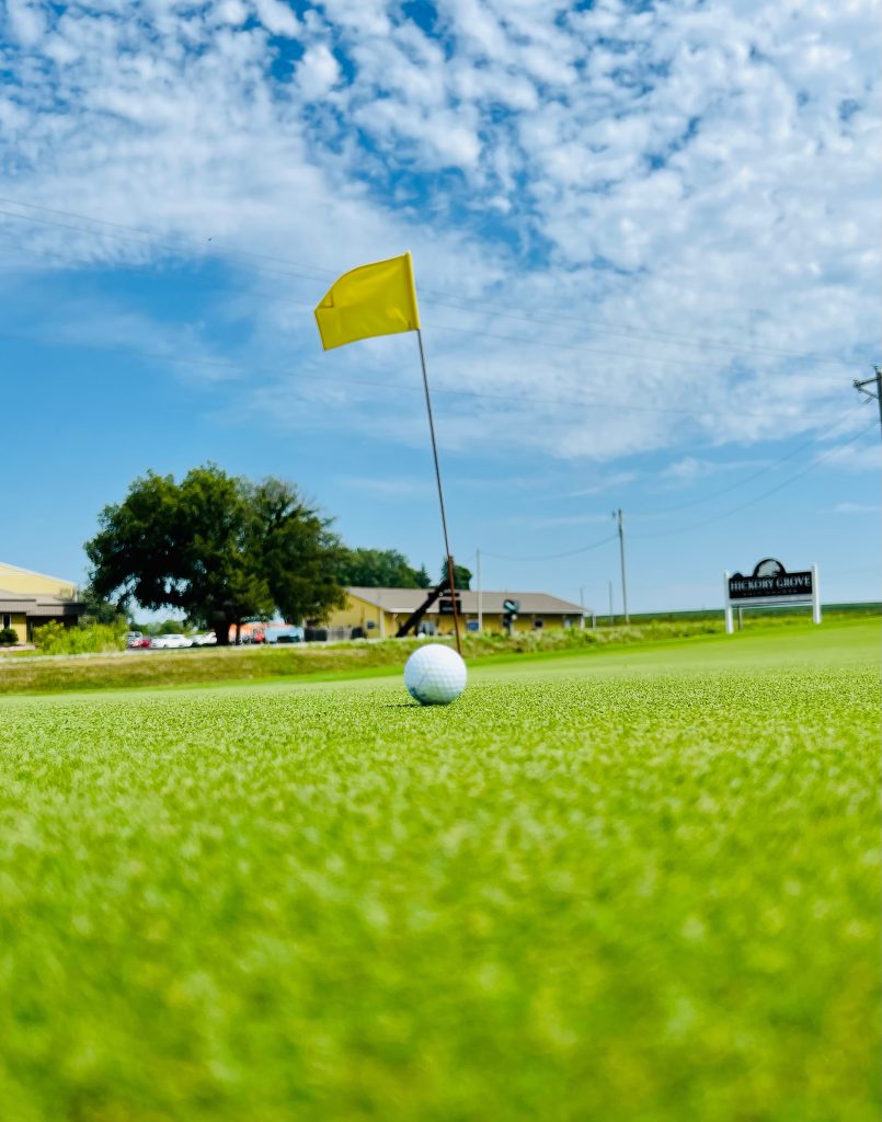 Golf ball on the green with a yellow flag in the hole.